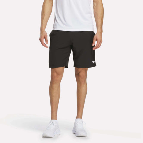 A person wearing a white shirt, black Reebok Men’s Range Shorts 9″, and white sneakers stands against a plain white background. Only the lower torso and legs are visible.
