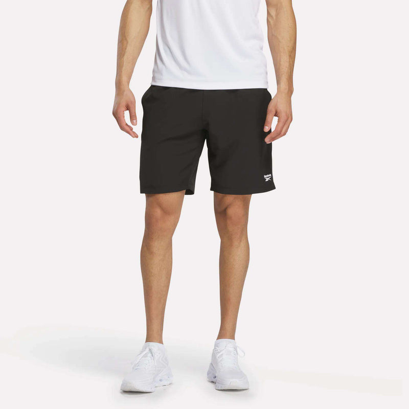 A person wearing a white shirt, black Reebok Men’s Range Shorts 9″, and white sneakers stands against a plain white background. Only the lower torso and legs are visible.