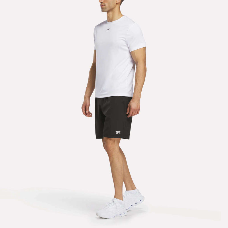 A man wearing a plain white T-shirt, Reebok Men’s Range Shorts 9″ in black, and white sneakers stands facing slightly to the side against a white background.