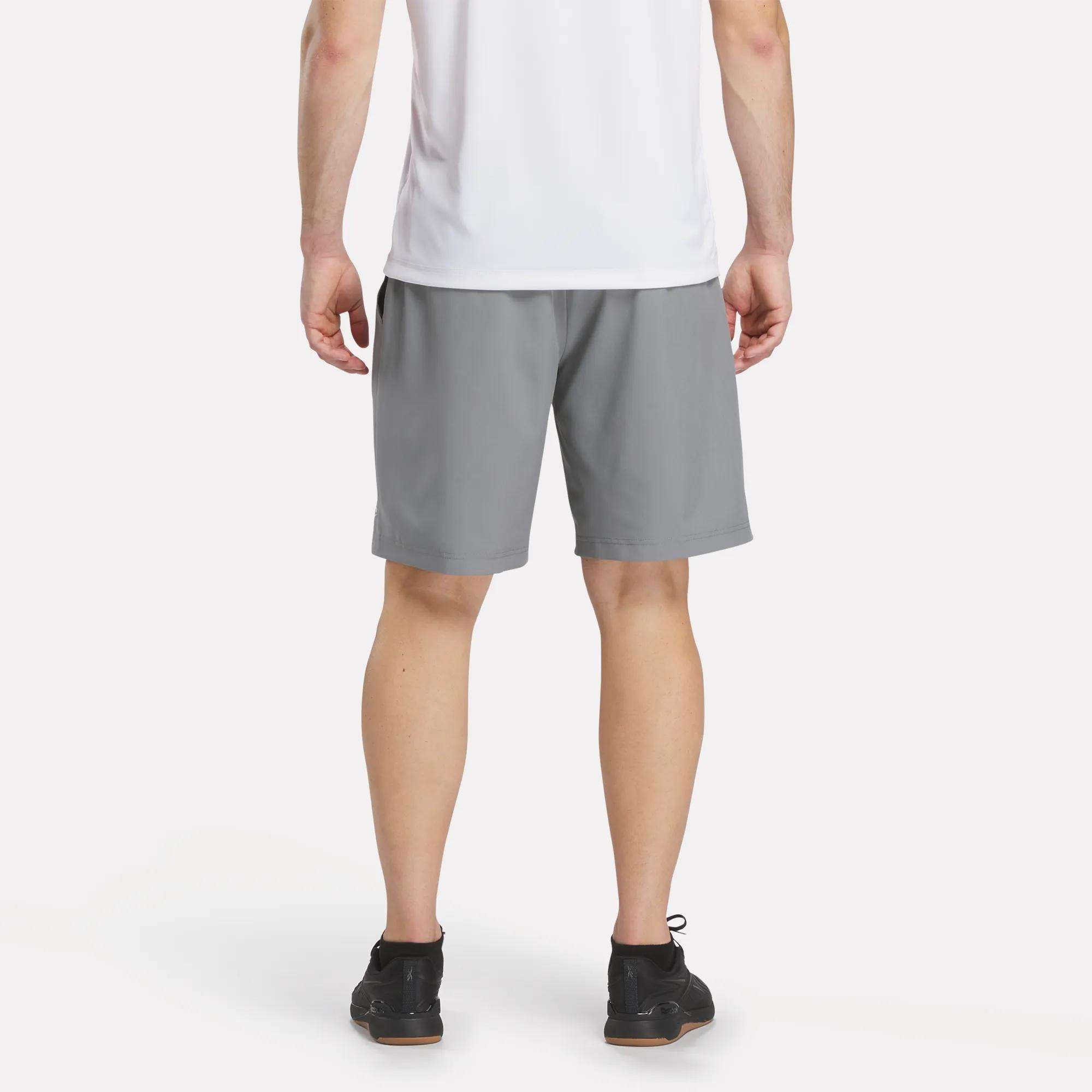 A person stands with their back to the camera, wearing a white t-shirt, Reebok Men's Range Shorts 9", and black sneakers against a plain white background.