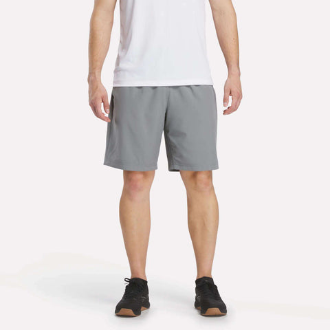 A man stands facing forward in a white t-shirt, Reebok Men's Range Shorts 9", and black sneakers against a plain light background. His head is not visible in the image.