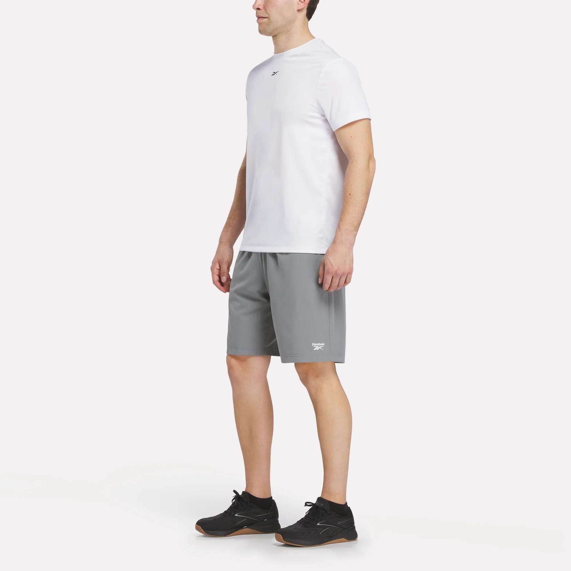 A man stands facing slightly left, wearing a white short-sleeve shirt, Reebok Men's Range Shorts 9" in gray, and black sneakers against a plain white background.