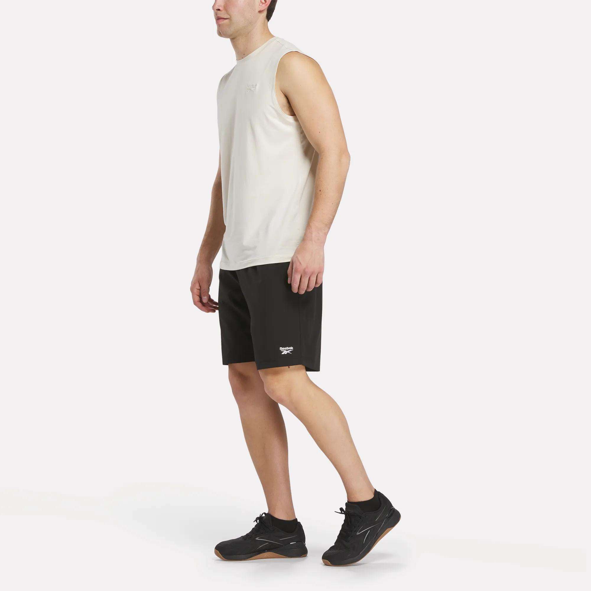 A man in a Reebok Men's Elite Sleeveless Top, paired with black athletic shorts and black sneakers, stands facing left against a plain white background.