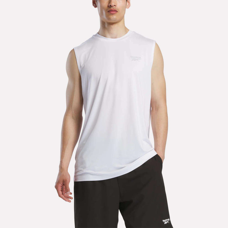 A man wears a Reebok Men's Elite Sleeveless Top in white cotton flex knit and black shorts, standing with one hand in his pocket against a plain background.