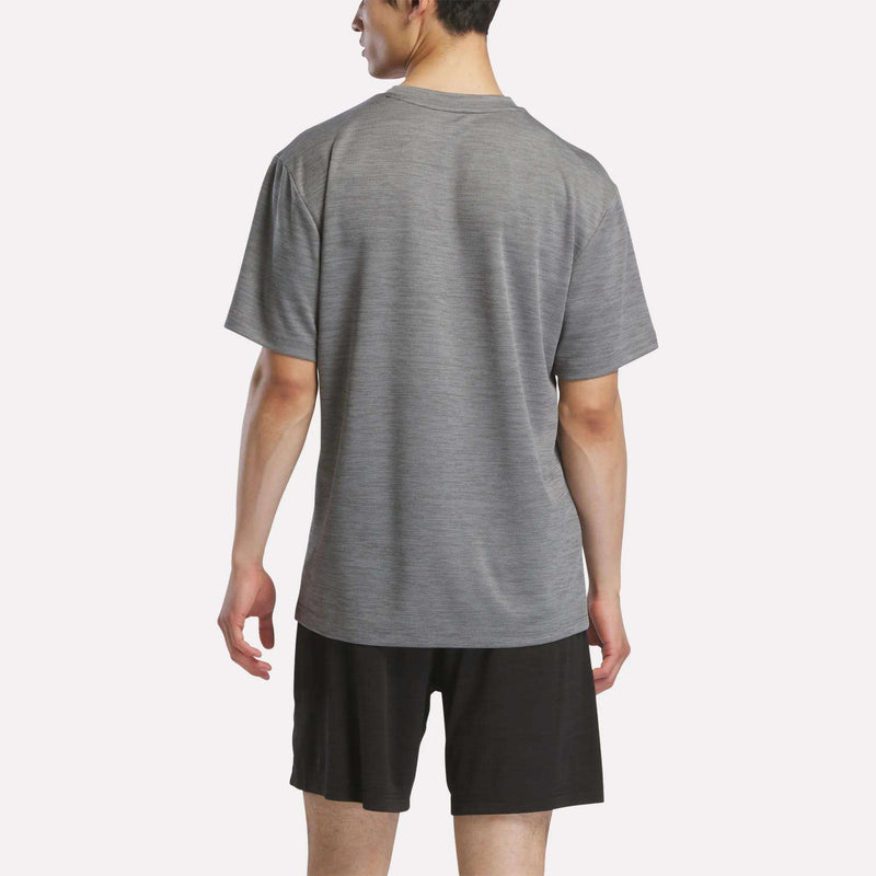 A person stands with their back to the camera, wearing a Reebok Men's Director Short Sleeve Top and black shorts against a plain white background.
