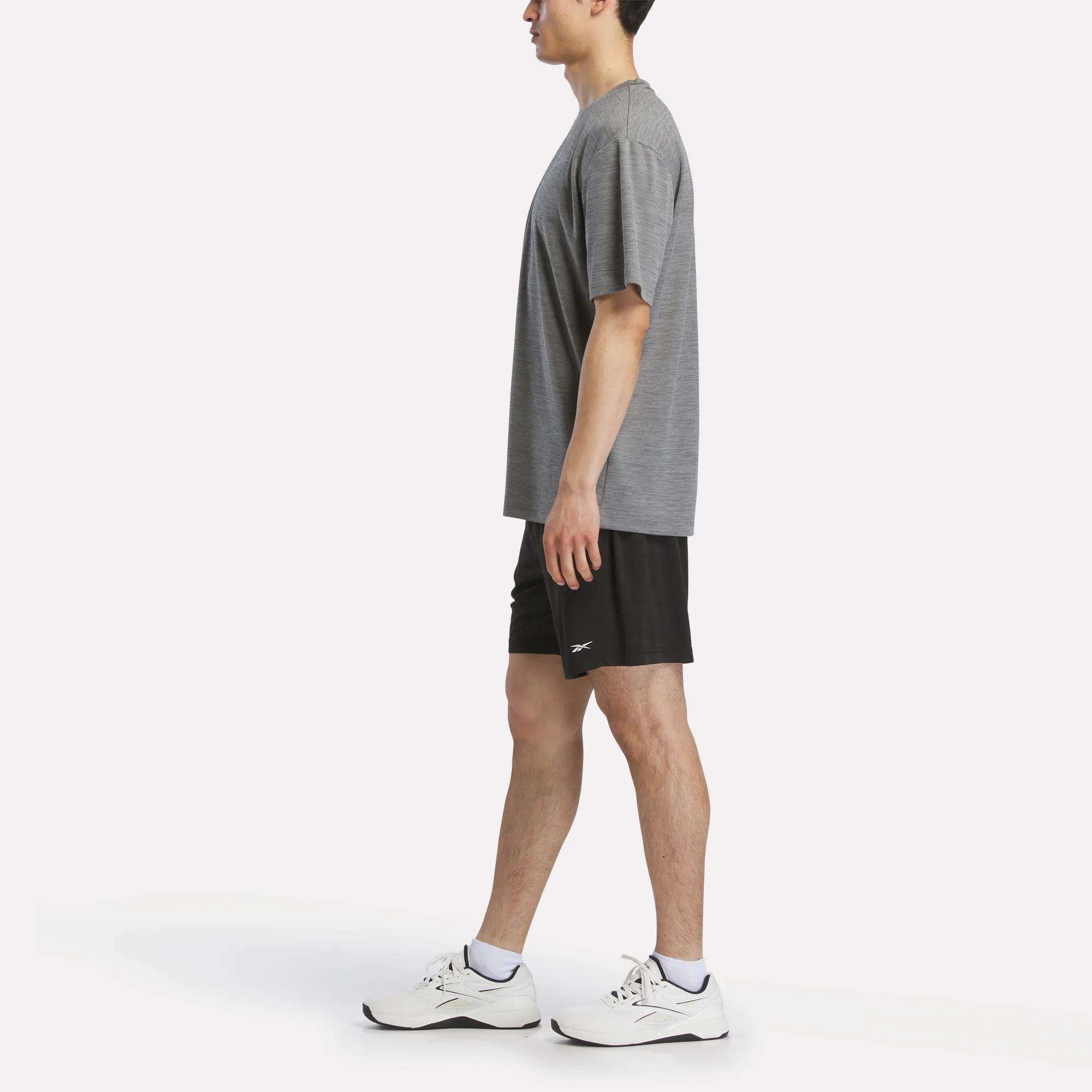 A man in a Reebok Men's Director Short Sleeve Top, black athletic shorts, and white sneakers stands in profile facing left against a plain white background.