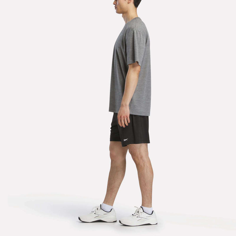 A man in a Reebok Men's Director Short Sleeve Top, black athletic shorts, and white sneakers stands in profile facing left against a plain white background.