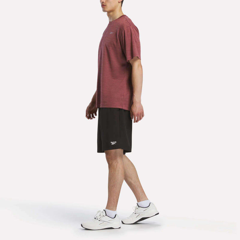 A person stands in profile against a plain white background, wearing a maroon Reebok Men's Director Short Sleeve Top, black shorts, white socks, and white athletic shoes.