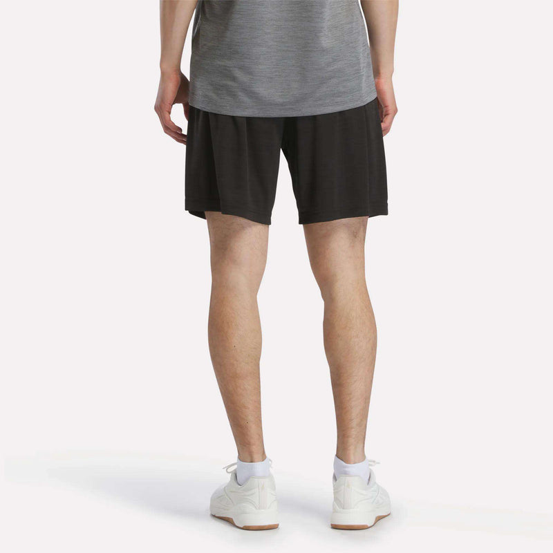 A person stands facing away, wearing a gray shirt, Reebok Men's Inclination Shorts in black, and white sneakers. The background is plain white.