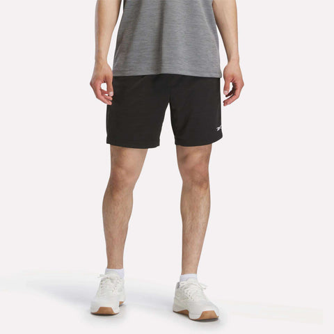 A person wearing a gray t-shirt, white sneakers, and Reebok Men's Inclination Shorts in black with a small white logo, standing against a plain light background.