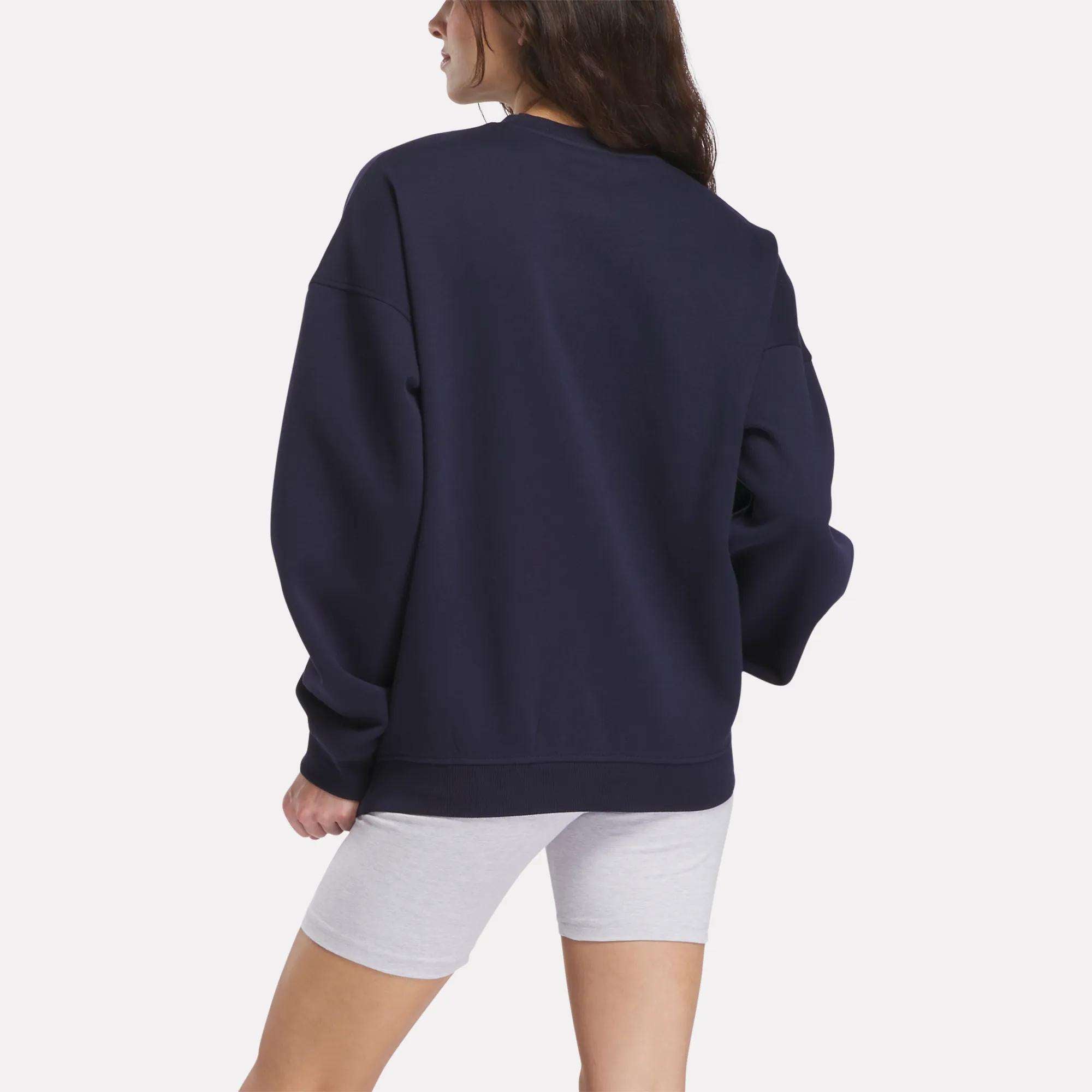 A woman with long brown hair stands with her back to the camera, wearing the Reebok Relaxed Fit Crew Neck Sweatshirt in navy blue and light gray fitted shorts, set against a plain white background.