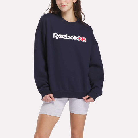 A person stands against a plain white background, wearing a Relaxed Fit Crew Neck Sweatshirt by Reebok in navy blue with a UK flag logo and light gray shorts, embodying classic heritage style with a relaxed fit.