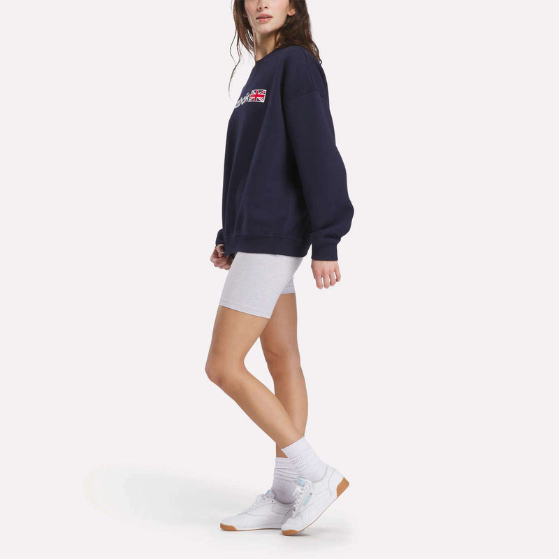 A woman poses against a plain background, wearing a navy Reebok Relaxed Fit Crew Neck Sweatshirt, light gray bike shorts, white crew socks, and white sneakers. She stands with her head slightly turned and her hands by her sides.