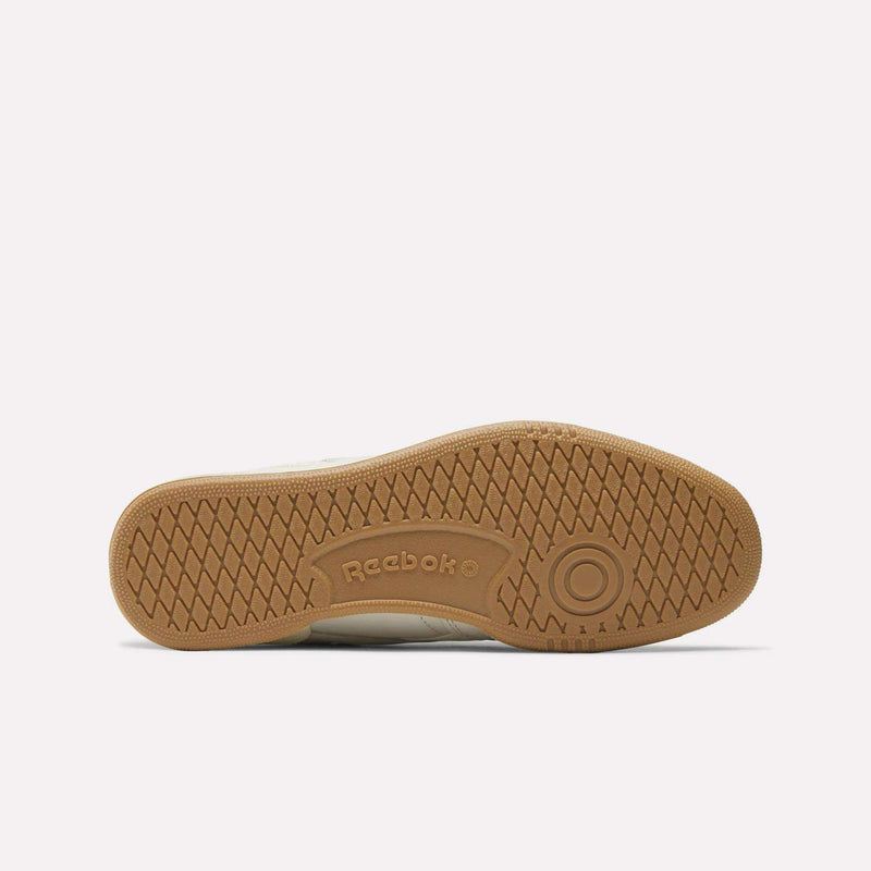 The image shows the gum-colored rubber outsole of the Reebok Men's Club C Revenge Shoes from below, highlighting its textured, patterned tread with the Reebok logo and a circular design near the heel.