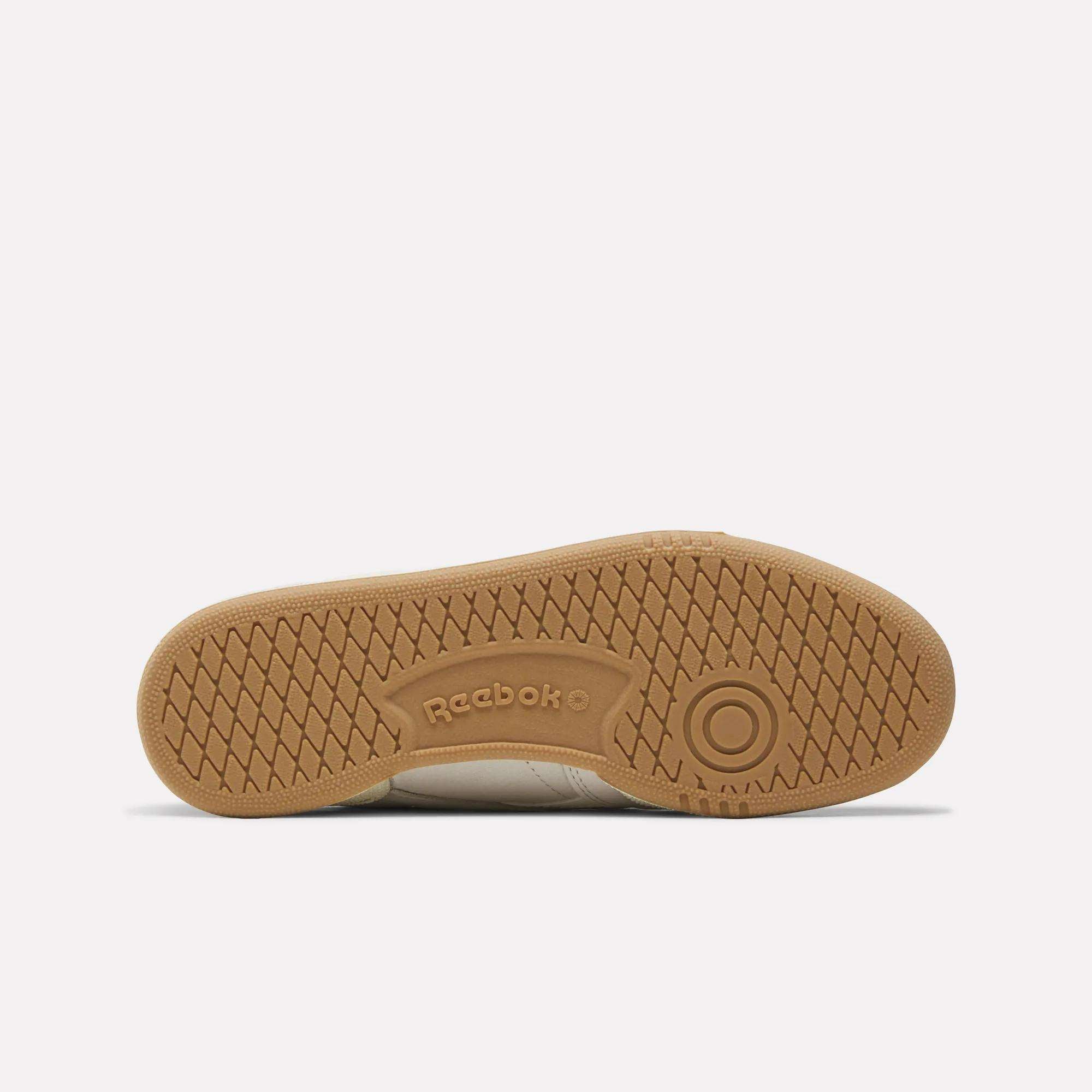 The image shows the sole of the Reebok Women's Club C Revenge Shoes, highlighting a light brown gum rubber outsole with a herringbone tread pattern and the Reebok logo embossed in the center.
