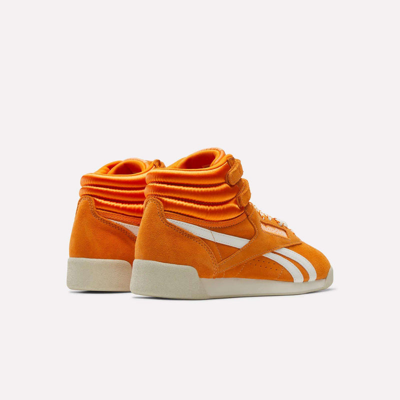 Women’s Freestyle Hi Shoes by Reebok in orange, showcasing a retro hi-top design with double Velcro ankle straps, white side stripes, and white soles, displayed against a plain white background.