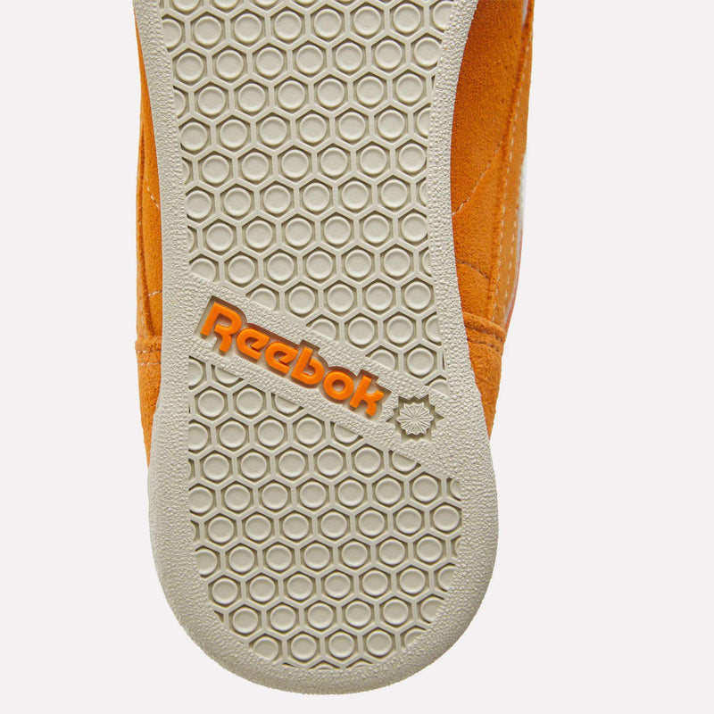 Close-up of the sole of a Reebok Women’s Freestyle Hi Shoe in orange, featuring a hexagonal tread pattern and the Reebok logo in orange lettering—an eye-catching detail on these women’s sneakers.