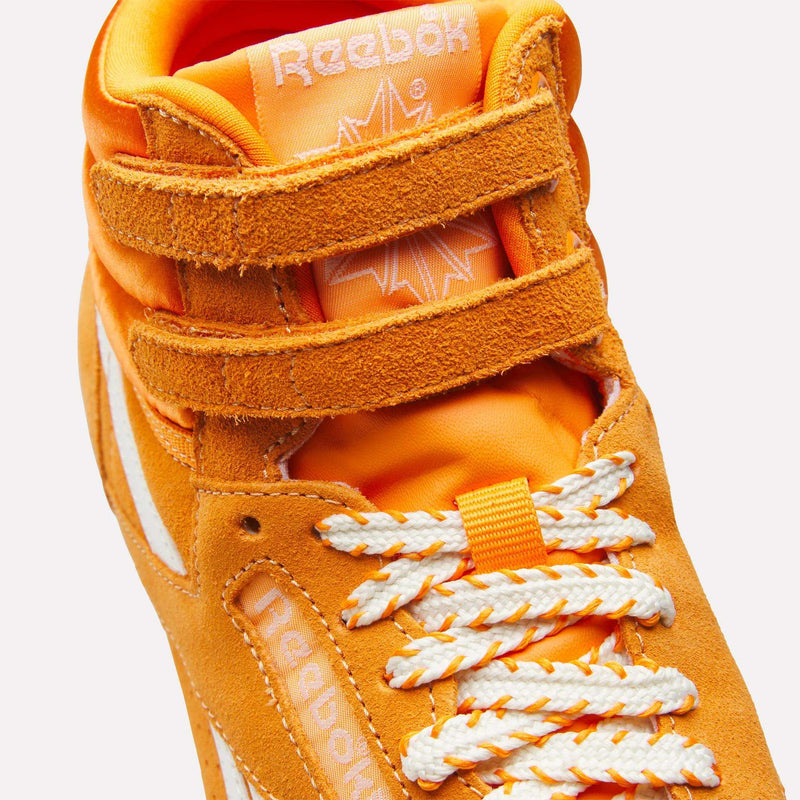 Close-up of the Reebok Women’s Freestyle Hi Shoes in orange, featuring laces, Velcro straps, and the Reebok logo on the tongue and side. The sneaker stands out with white accents and textured suede material, making it a distinctive choice among women’s footwear.