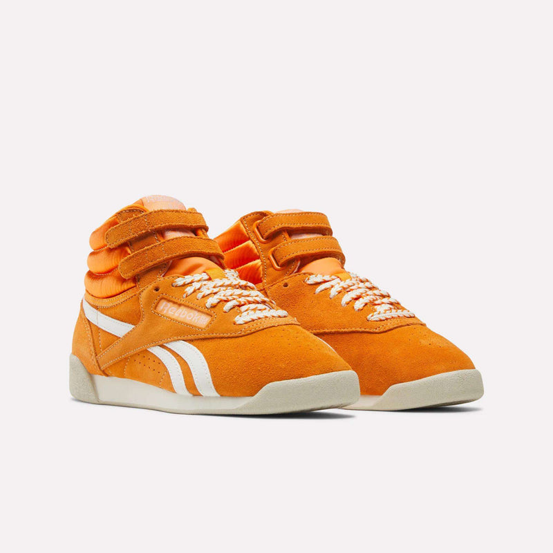 A pair of Women’s Freestyle Hi Shoes by Reebok in bright orange, featuring white stripes, velcro straps, patterned white laces, and beige soles, displayed against a plain white background. These retro sneakers embody classic Reebok style with their eye-catching color and iconic hi-top silhouette.