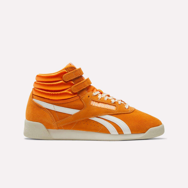 The Women’s Freestyle Hi Shoes by Reebok feature a bright orange hi-top design with white accents, two velcro straps, orange-and-white laces, and a white sole, shown against a plain white background.