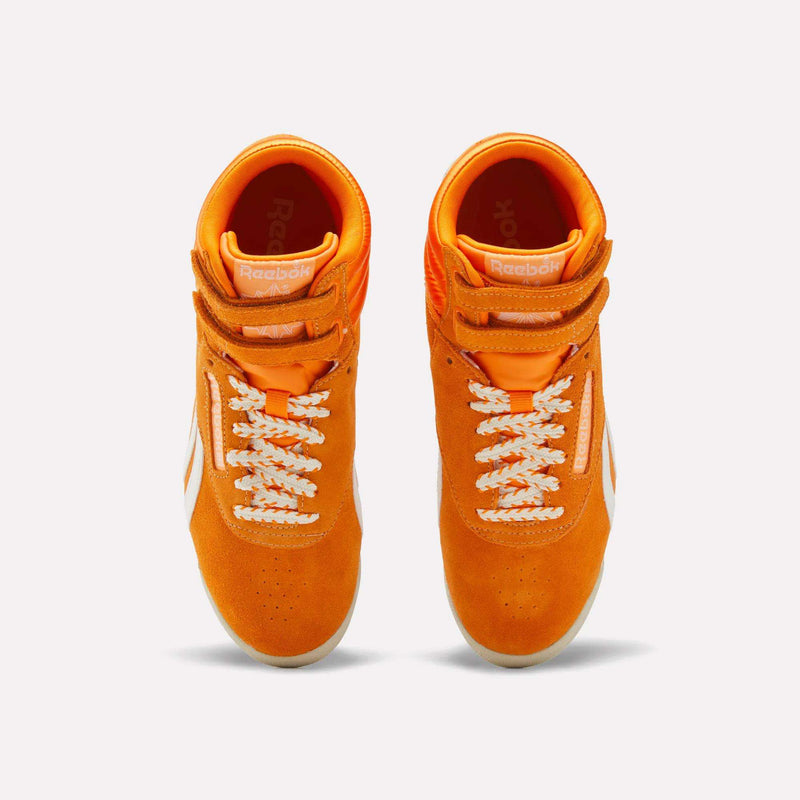 Top view of a pair of Reebok Women’s Freestyle Hi Shoes in bright orange, featuring white laces and side stripes, placed side by side on a white background.