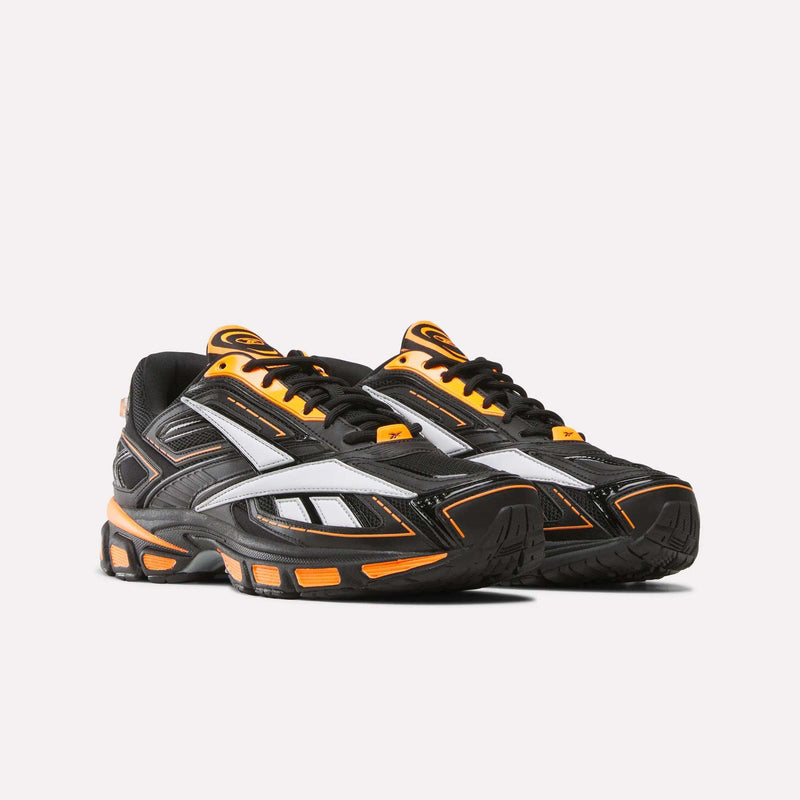 A pair of Reebok Premier Road Ultra SV Running Shoes in black with orange and white accents, featuring a bold white geometric design on the sides and thick black soles highlighted with orange details, evoke retro Y2K sneaker aesthetics inspired by the iconic Premier Road Ultra style—all presented against a plain light background.
