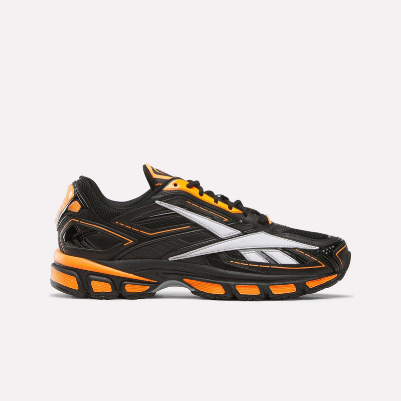 The Reebok Premier Road Ultra SV Running Shoes feature a black base with orange and white accents, including orange details on the sole and upper. A bold white Reebok logo stands out on the side, channeling retro Y2K sneaker style. Shown against a plain white background.