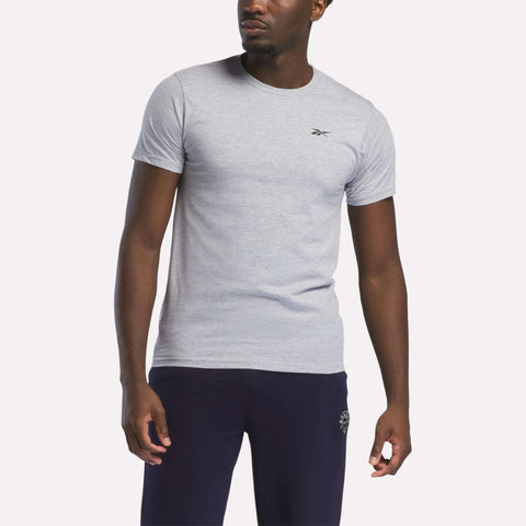 A man in a relaxed fit light gray Reebok Men's Small Logo T-Shirt and dark pants stands against a plain white background.