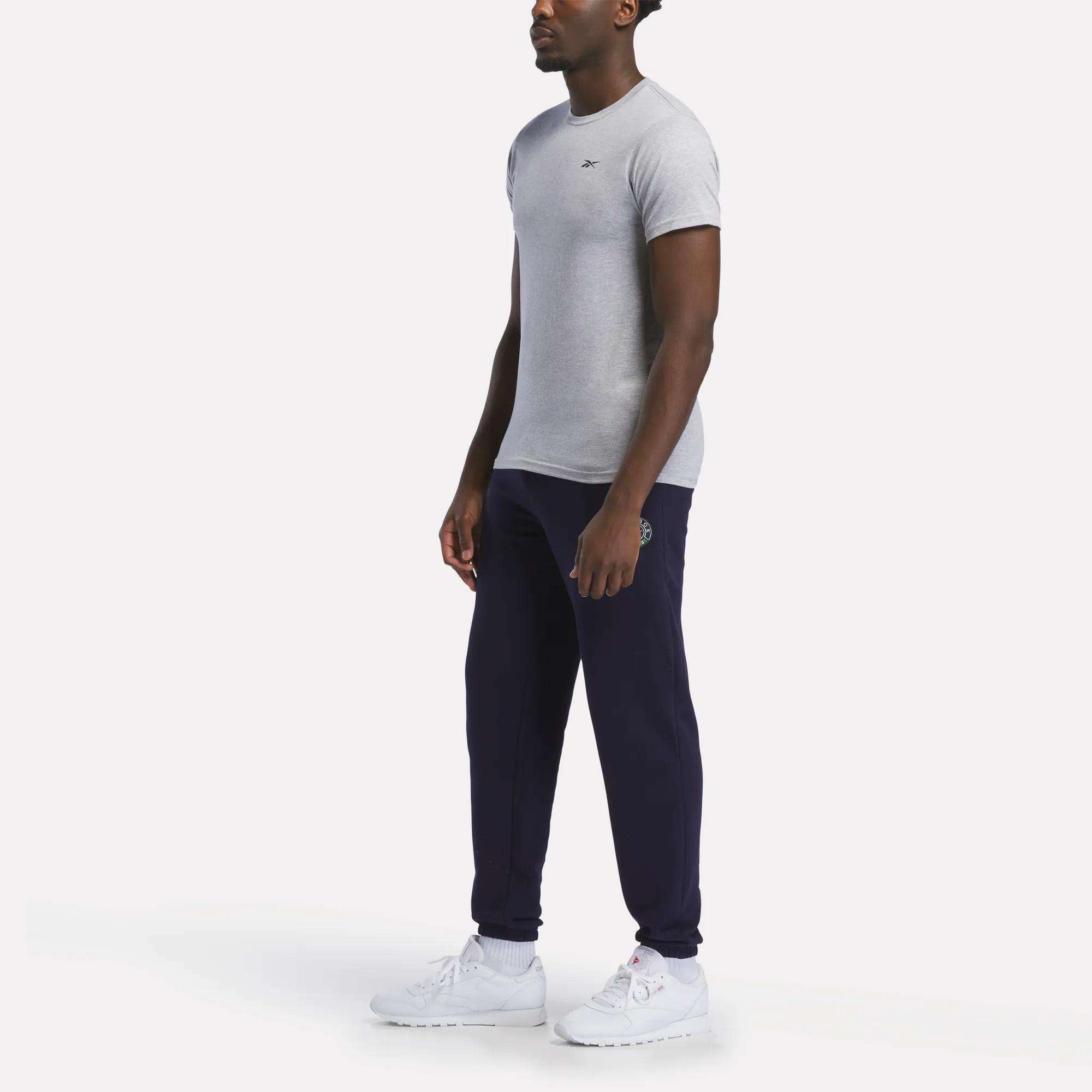 A man stands against a plain white background wearing a relaxed fit Reebok Men's Small Logo T-Shirt in grey, dark blue jogger pants, and white sneakers.