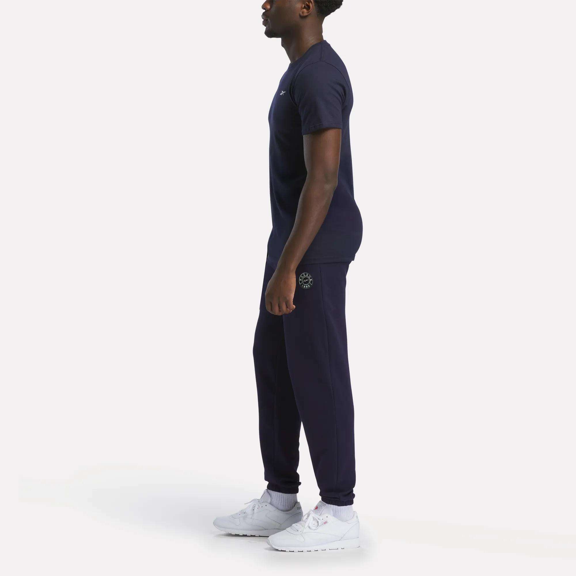 A person stands in profile wearing a relaxed fit Reebok Men's Small Logo T-Shirt in navy blue with joggers and white athletic shoes, set against a plain white background.