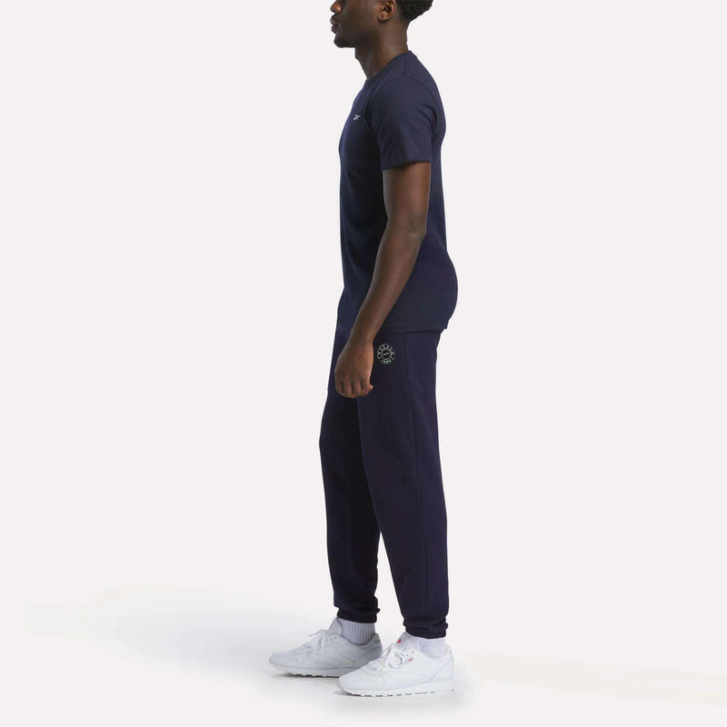 A person stands in profile wearing a relaxed fit Reebok Men's Small Logo T-Shirt in navy blue with joggers and white athletic shoes, set against a plain white background.