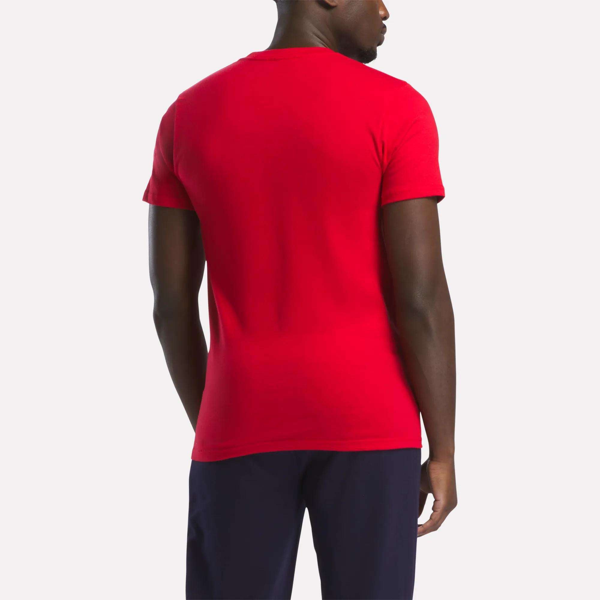 A man is seen from the back in a relaxed fit red Reebok Men's Small Logo T-Shirt and dark pants, standing against a light gray background.