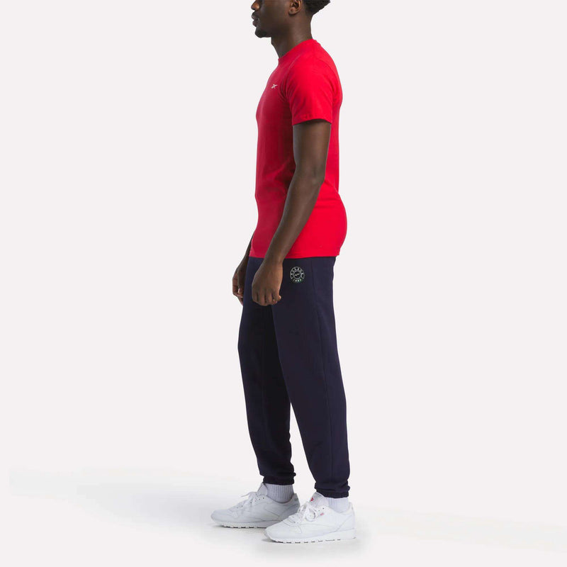 A person stands in profile against a white background, wearing a relaxed fit Reebok Men's Small Logo T-Shirt, navy jogger pants with a small logo on the hip, and white sneakers.