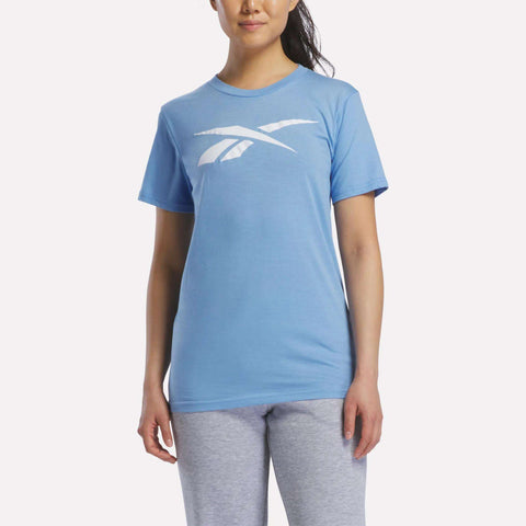 A woman stands against a plain white background, wearing a light blue Reebok Women's Vector Graphic T-Shirt with a bold white logo on the chest, paired with light gray pants.