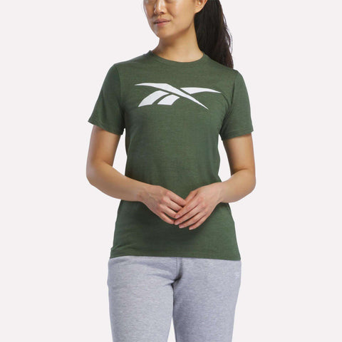 A woman wears a Reebok Women's Vector Graphic T-Shirt in dark green with a large white logo, paired with light gray sweatpants, standing against a plain white background.
