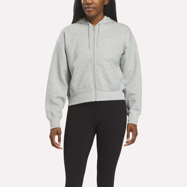 A person wears a Reebok Women's Cropped Full-Zip Hoodie in Grey Heather, featuring front pockets, paired with black leggings against a plain light background.