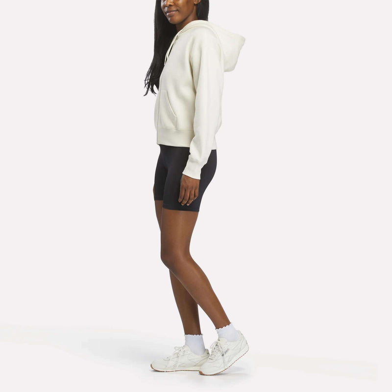 A woman stands sideways against a white background, wearing a Reebok Women's Cropped Full-Zip Hoodie, black biker shorts, white socks, and white sneakers.