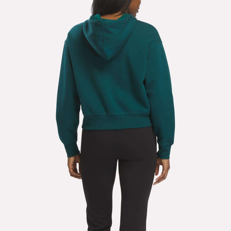 A woman is seen from the back wearing a teal Reebok Women's Cropped Full-Zip Hoodie with an embroidered logo and black leggings, standing against a plain white background.