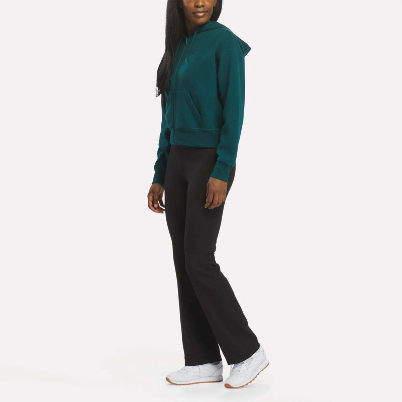 A woman in a teal Reebok Women's Cropped Full-Zip Hoodie, black pants, and white sneakers stands against a plain white background, turned slightly to the side.