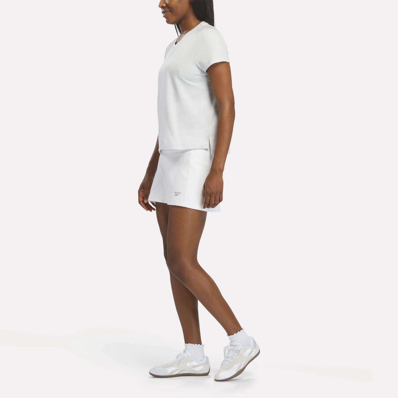 A woman faces left against a plain white background, wearing the Reebok Women's Graphic Glow Top with a matching skirt, paired with white athletic shoes and socks.
