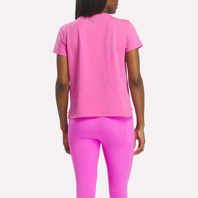 A woman stands with her back to the camera, wearing the Reebok Women's Graphic Glow Top in bright pink and matching leggings. The embroidered logo is visible, set against a plain light background.