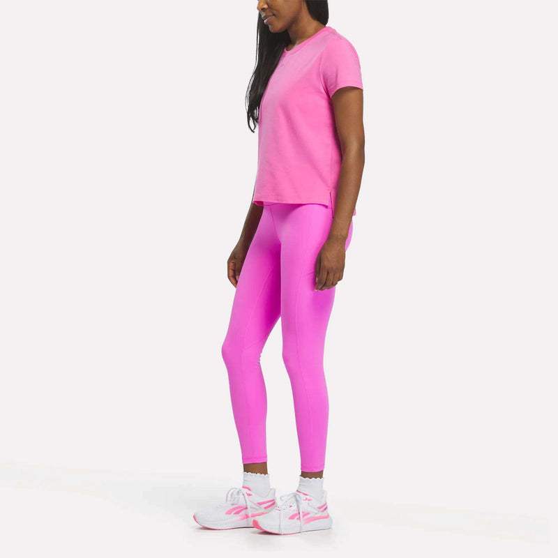 A woman stands sideways against a plain white background, wearing a bright pink Reebok Women's Graphic Glow Top, matching pink leggings, and white sneakers with pink accents.