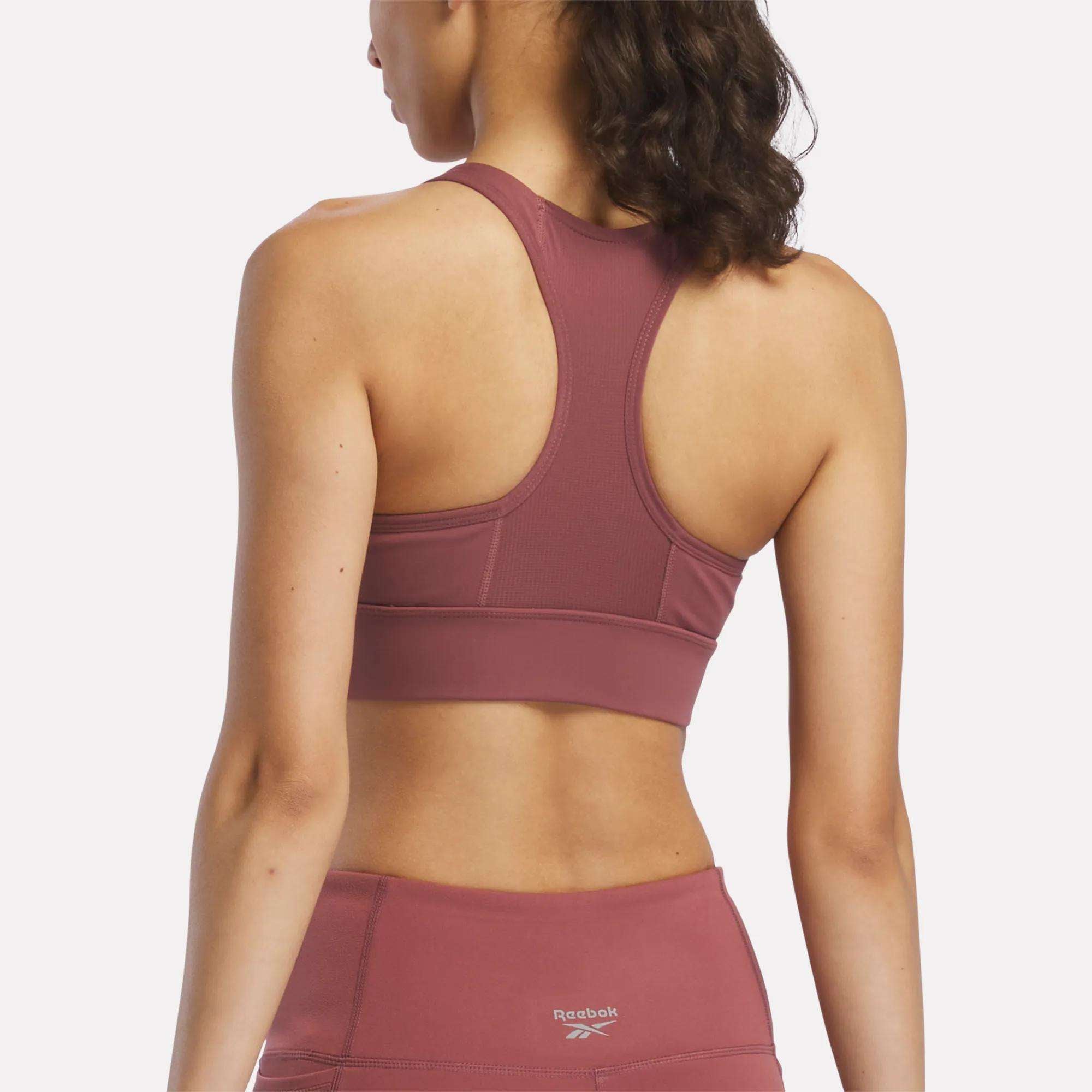 A woman with wavy brown hair wears the Reebok Women's Rush Medium-Impact Sports Bra and matching mauve leggings, shown from the back against a plain white background. The outfit is ideal for medium-impact workouts.
