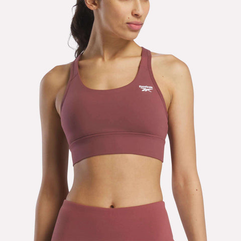 A woman in a mauve Reebok Women's Rush Medium-Impact Sports Bra and matching high-waisted leggings poses against a plain, light background, showcasing activewear suited for medium-impact workouts.