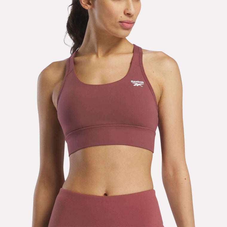 A woman in a mauve Reebok Women's Rush Medium-Impact Sports Bra and matching high-waisted leggings poses against a plain, light background, showcasing activewear suited for medium-impact workouts.