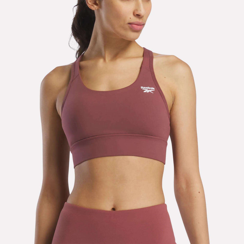 A woman in a mauve Reebok Women's Rush Medium-Impact Sports Bra and matching high-waisted leggings poses against a plain, light background, showcasing activewear suited for medium-impact workouts.