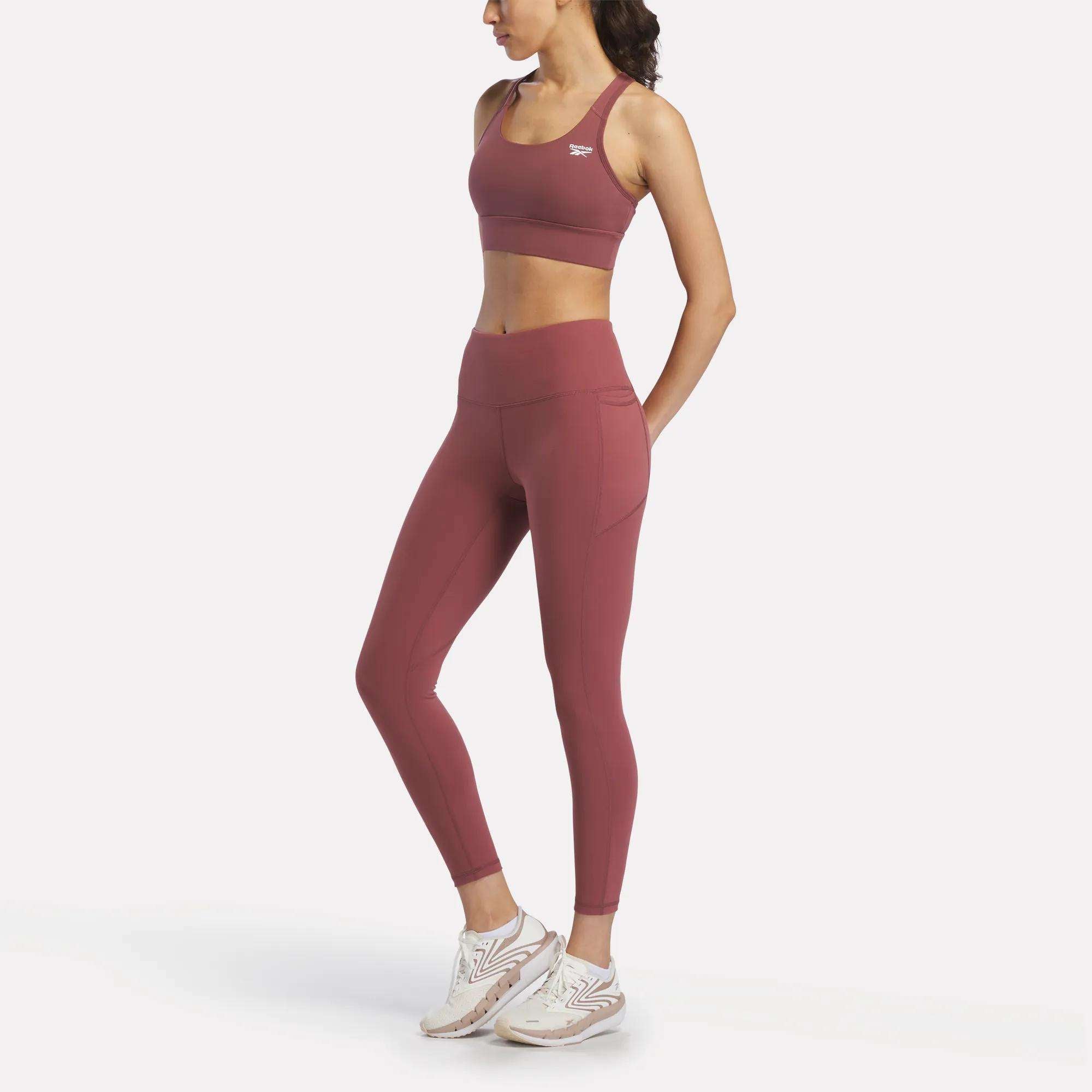 A woman in a matching Reebok Women's Rush Medium-Impact Sports Bra and high-waisted leggings with white sneakers stands relaxed against a plain white background, ideal for medium-impact workouts.