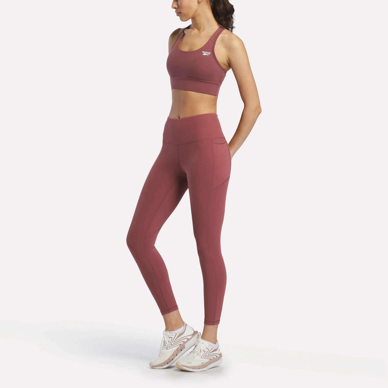 A woman in a matching Reebok Women's Rush Medium-Impact Sports Bra and high-waisted leggings with white sneakers stands relaxed against a plain white background, ideal for medium-impact workouts.