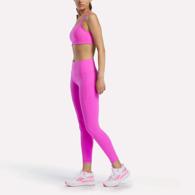 A woman stands sideways against a white background, wearing a bright pink sports bra, Reebok Women's Odyssey High-Rise 7/8 Leggings, and white and pink athletic shoes.