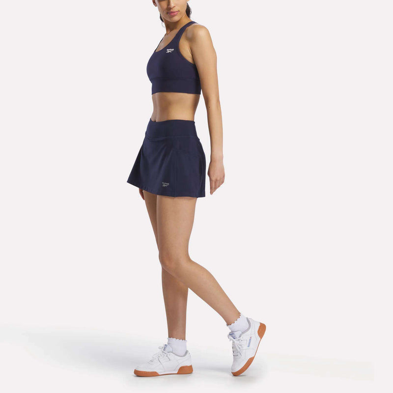 A woman poses against a plain white background wearing the Reebok Women's Break Point Skort in navy blue with a matching sports bra, white socks, and white sneakers with brown soles.